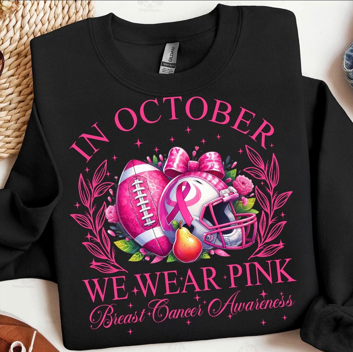 A black crewneck sweatshirt featuring a pink and multicolor graphic design for Breast Cancer Awareness. The graphic includes the text 'IN OCTOBER WE WEAR PINK' in pink lettering, surrounding an illustration of a pink football, a pink-and-white helmet with a ribbon, a pink bow, and a pear.