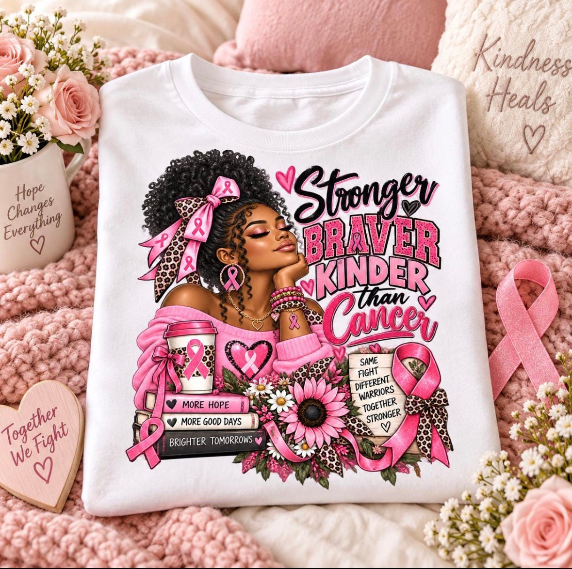 A white graphic T-shirt laid flat on a pink knitted blanket with roses, ribbons, and heart-shaped signs around it. The shirt features a large printed illustration of a Black woman with an afro, pink bow, and pink awareness ribbons, alongside the text 'Stronger Braver Kinder than Cancer' and breast cancer awareness motifs.