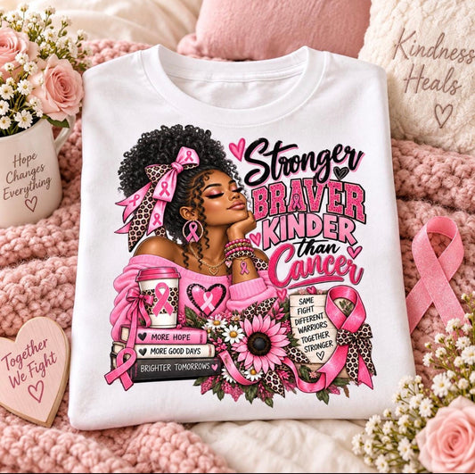 A white graphic T-shirt laid flat on a pink knitted blanket with roses, ribbons, and heart-shaped signs around it. The shirt features a large printed illustration of a Black woman with an afro, pink bow, and pink awareness ribbons, alongside the text 'Stronger Braver Kinder than Cancer' and breast cancer awareness motifs.